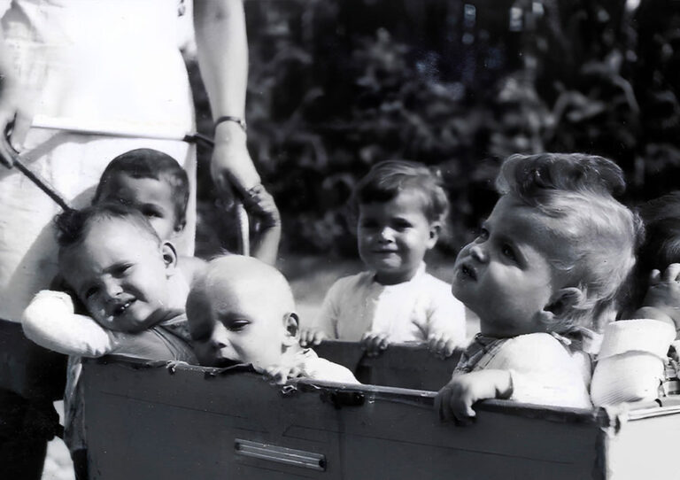 Grup de nadons i nens petits en un entorn exterior, probablement un jardí o un parc. Els nens estan asseguts o dempeus dins d'una caixa o carro de fusta.