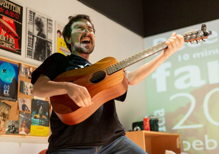 Home tocant una guitarra acústica en un espai interior. L'home està cantant amb passió, amb la boca oberta i els ulls tancats. Porta una samarreta negra i texans blaus.
