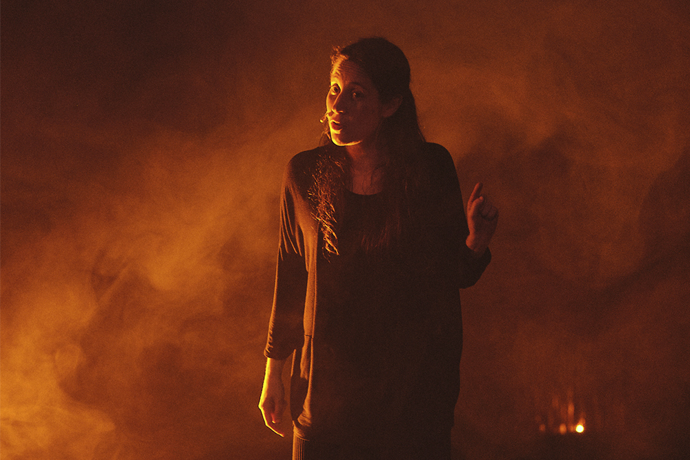 Una dona en un escenari teatral, vestida amb un vestit negre de màniga llarga. Està mirant cap a l'esquerra de la imatge i té la mà dreta aixecada.