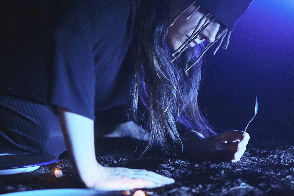 Persona agenollada a terra, amb el cap cot i els cabells llargs i foscos que li cobreixen la cara. Està vestida amb roba fosca i sosté una cullera a la mà.