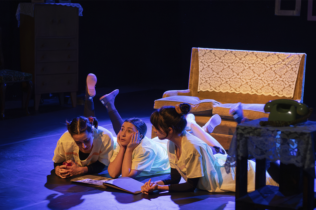 Three women lying on the floor in a dark scenario with orange sofa and green telephone, watching a book.