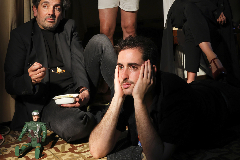 A man with a dark jacket and dark t -shirt holds a fork with food and a lunch box. Another man, with a dark t -shirt, rests his head in his hands. In the background, you can see a person's legs with white shorts and a person's feet with heels. On the floor, in front of the man in the jacket, is a figure of soldier action.