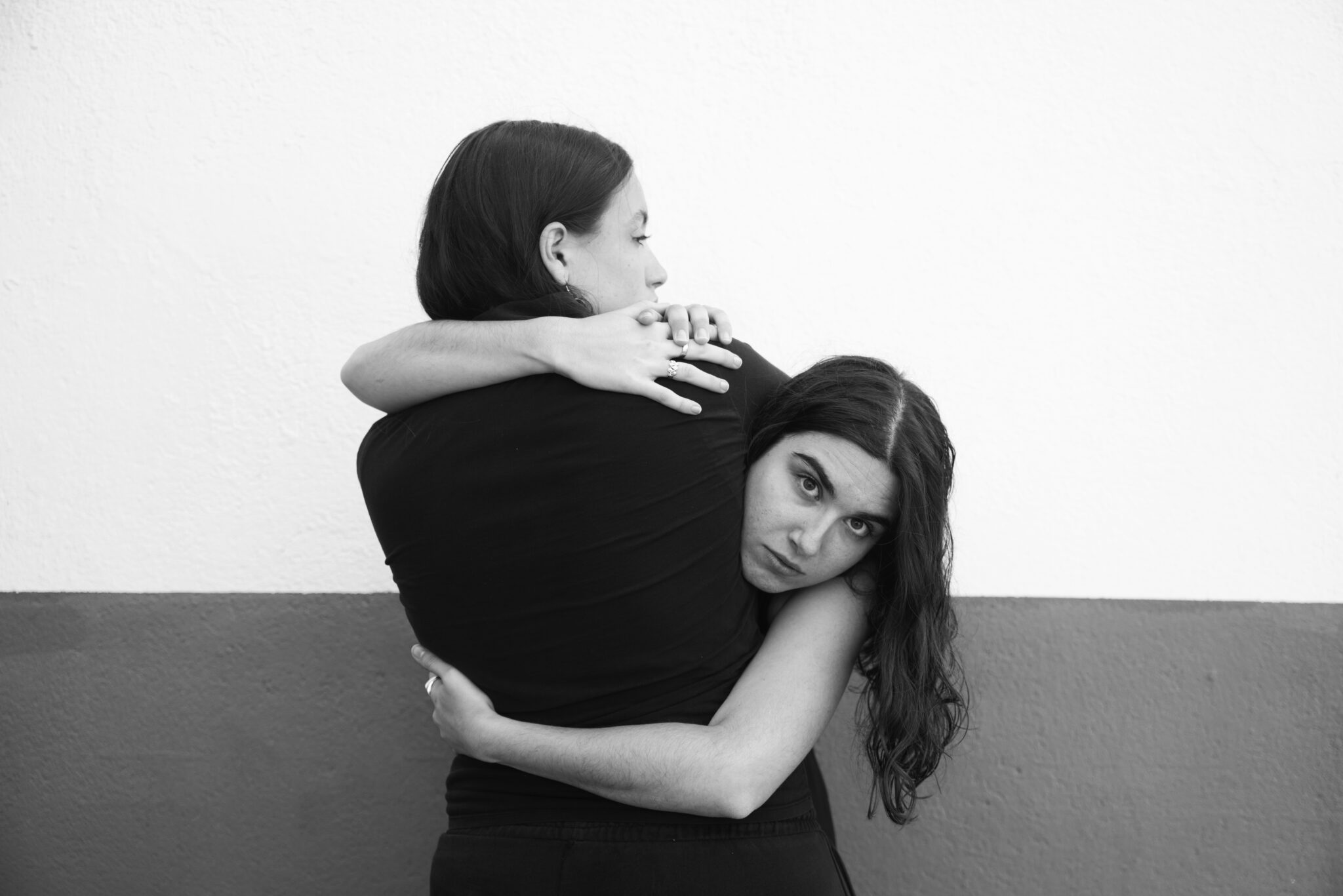 A black and white photograph of two women hugged, with one of them looking directly on the camera while the other looks sideways, both in dark clothes.