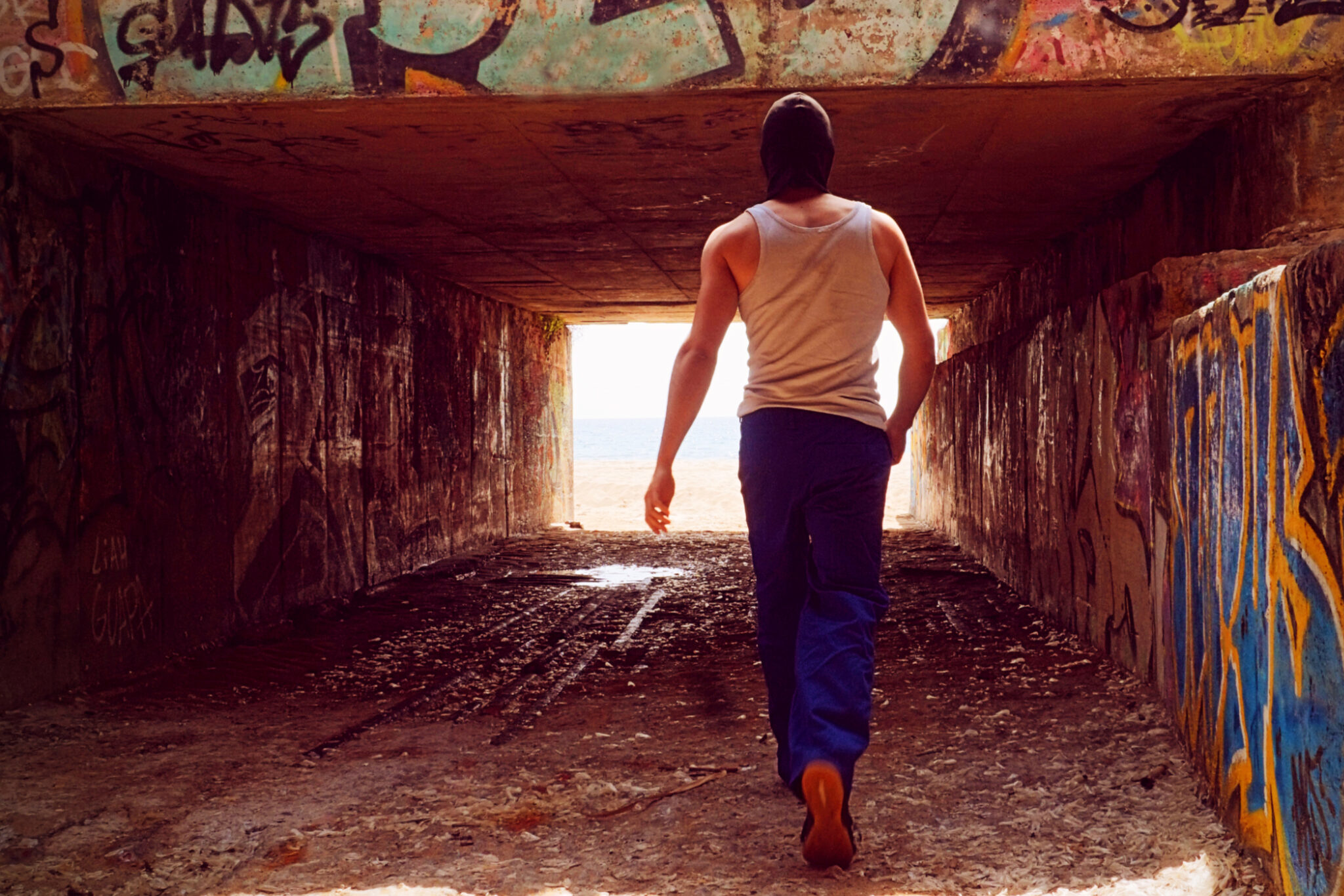 A back guy dressed in blue work pants and t -shirt. He carries a handrail and is about to cross under a bridge. The bridge is full of graffiti.
