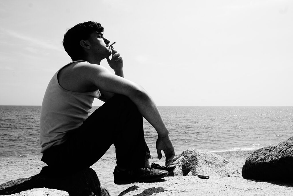 Una fotografia en blanc i negre d'un home assegut en unes roques a la vora del mar, amb un cigarret a la mà, mirant cap al cel.