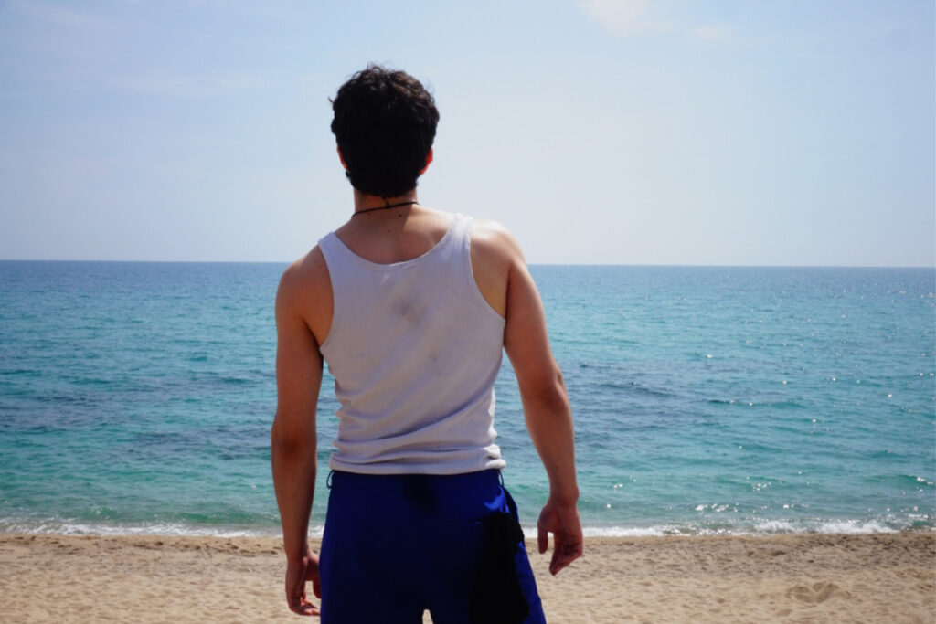 Una fotografia d'esquena d'un home dret a la platja, amb samarreta de tirants blanca i pantalons blaus, mirant cap al mar sota un cel clar.