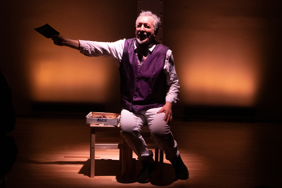 Un home de cabells blancs, vestits de manera elegant. Està assegut en un banc de fusta. En un espai teatral. Darrere, un fons de color taronja.