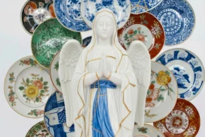 Una estàtua de porcellana blanca de la Mare de Déu amb ales, amb les mans juntes en oració, envoltada per una col·lecció de plats de ceràmica decoratius de diversos estils.